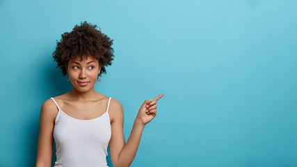 Portrait of lovely curly haired Afro American woman points at copy space and advertises something...