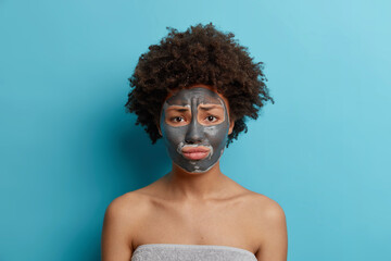 Photo of worried curly haired woman applies nourishing charcoal mask on face wants to have perfect smooth skin stands wrapped in towel after taking bath isolated over blue studio background.