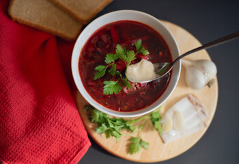 Traditional Ukrainian Russian borscht or red soup with a drop of fresh sour cream and with a fresh sprig of green parsley in the white bowl. Lard, garlic, bread and fresh herbs for soup near. 