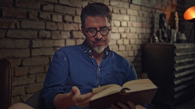 Mature man in his 50s reading book at home sitting in dark living room. 