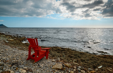Fototapeta na wymiar Red wooden armchair on a stone wild beach against the background of the sea and sky, the sun is hidden in the clouds. Seaside, work during lockout, copy space