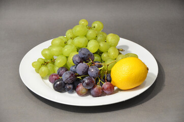 Healthy food. White plate with fruit. Vitamin breakfast. White and blue grapes on a gray background. Yellow lemon with grapes.