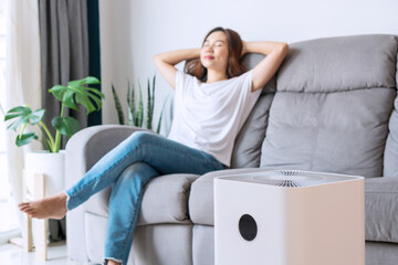 Pretty young Asian woman on comfortable sofa, hands behind head rest at home wellbeing breathing fresh air from air purifier while dust air pollution situation outside is r
