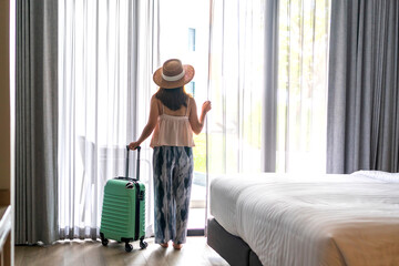 Young Asian traveler woman wearing straw hat and looking outside the window with her luggage in hotel room after check-in. Travel and vacation concept.