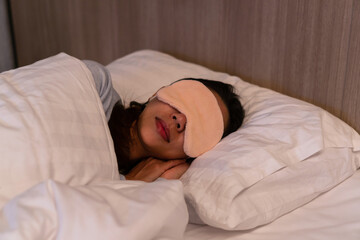 Closeup of young Asian female in sleeping mask lying in comfortable white bed at night.