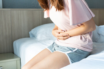 Fototapeta na wymiar Unhealthy young woman sitting on bed and holding belly, feeling discomfort and suffering from stomachache, food poisoning, on period. Health problem concept