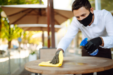 Waiter wearing protective face mask and gloves while disinfecting tables at outdoor cafe. Cleaning...