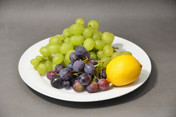 Multi-colored grapes and yellow lemon laying on a white plate. Fruit on a gray background.