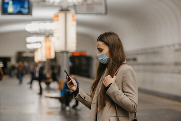 A woman in a face mask to avoid the spread of coronavirus is using a smartphone at a subway station. A girl in a surgical face mask against COVID-19 is waiting for a train on a metro platform.