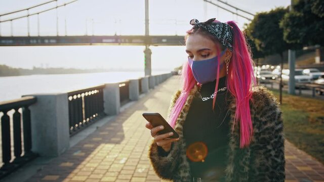 Young hipster lady in informal outfit and protective mask is using smartphone while standing at embankment