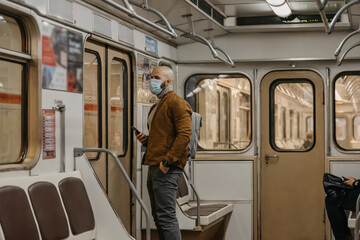 A man with a beard in a face mask to avoid the spread of coronavirus is waiting for a new stop in a subway car. A bald guy in a surgical mask against COVID-19 is holding a cellphone on a metro train.