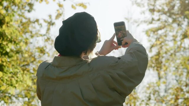 the back of a young woman takes a selfie photo or video in autumn against the background of trees and autumn leaves