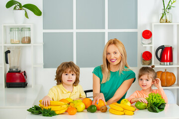 Happy laughing children and her beautiful young mother making fresh strawberry and other fruit juice for breakfast together in white kitchen with a window,