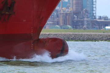 Bulbous bow of a ship with wave breaking before it as it passes industry of European port