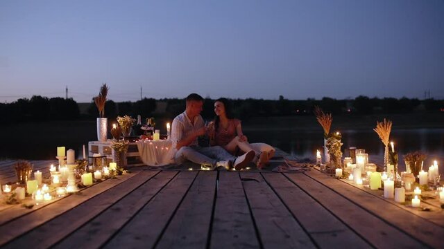 Romantic couple. Young people in love drink champagne on a deck decorated with candles. Happy couple sitting on a wooden deck. Marriage proposal concept. Romantic couple. Slow video.