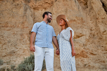 Young attractive couple on the background of rocks are holding hands and having fun. A woman in a hat and dress and a man in a shirt and white trousers are resting.