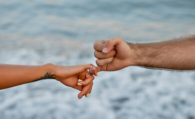 Close-up cropped image of a young couple's hands in front of the sea and waves, holding their little fingers in a gesture of acceptance.