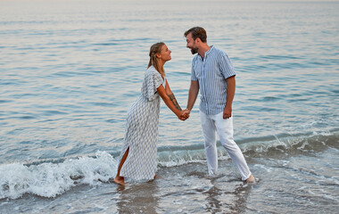 An attractive woman in a dress and a handsome bearded man in a striped shirt hold hands and romantically spend time on the seashore. A loving couple on vacation.