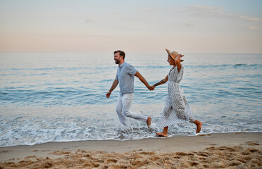 An attractive woman in a dress and a handsome bearded man in a striped shirt run holding hands and romantically spend time on the seashore. A loving couple on vacation.