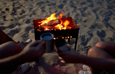 Cropped image of a man and woman holding mugs of tea by the fire, on a sandy beach by the sea.