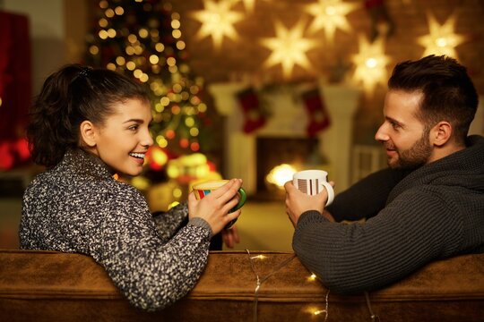 Christmas couple at home in Winter. Happy young couple drinking tea at home in Christmas time. Christmas tree and fireplace in background.