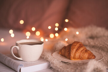 Cup of fresh tea staying on open paper book with knitted cloth and croissant in bed close up. Good morning. Breakfast time. Winter holiday season.
