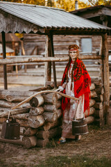A beautiful Slavic girl with long blonde hair and brown eyes in a white and red embroidered suit is sitting next to a wooden house.Traditional clothing of the Ukrainian region.