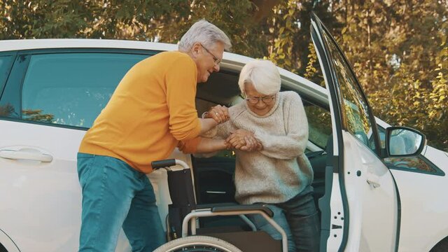 Elderly couple traveling. Man helping woman to get into the wheelchair from the car. High quality 4k footage