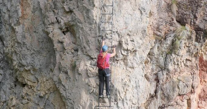Woman with red backpack climbs a vertical ladder on a via ferrata route called Piatra Soimilor, in Harghita county, Romania. Adventure, climbing, tilt up shot.