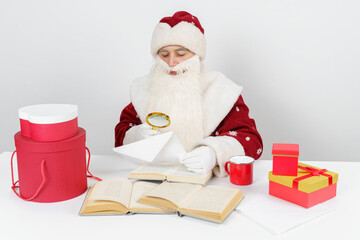 Tired Santa Claus sits at the table and examines letters with a magnifying glass. There are Christmas gifts and books on the table.