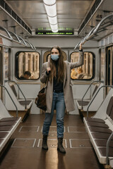 Fototapeta na wymiar A woman in a face mask to avoid the spread of coronavirus is holding onto the handrail in a subway car. A girl with long hair in a surgical mask against COVID-19 is standing on a metro train.