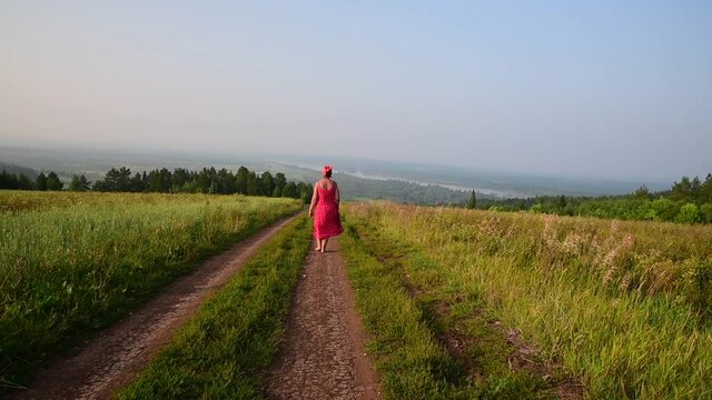 женщина в красном платье идет по полевой дороге на фоне красивого летнего  пейзажа