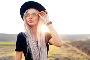 Portrait of young beautiful blonde girl, wearing black hat and yellow glasses on background of sunset.