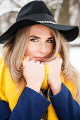 Outdoors lifestyle portrait of beautiful blonde girl smiling and walking in the snowy park. Wearing stylish dark blue coat, black hat, sunglasses and mustard scarf. Bright colors. Poitive emotions.