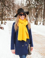 Outdoors lifestyle portrait of beautiful blonde girl smiling and walking in the snowy park. Wearing stylish dark blue coat, black hat, sunglasses and mustard scarf. Bright colors. Poitive emotions.