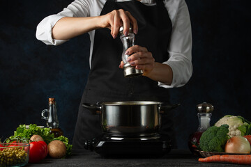 Close-up view of the professional chef in black apron pouring ground pepper into pot with soup on dark blue background. Backstage of cooking dinner. Preparing meal concept. Frozen motion.