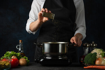 The professional chef in black apron adds spices into pot with soup on dark blue background....