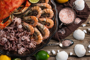 Close-up view of big dish with cooked crab, baby octopuses and tiger shrimps served with sea salt, lemon, lime and white seashells on rustic wooden background. Seafood concept. Delicious food.