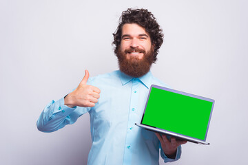 Portrait of young bearded man in casual showing thumb up gesture and holding laptop with green screen