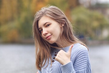 Portrait of young happy positive woman, teenager girl is showing thumb up, like, approval gesture, looking at camera and smiling in autumn park