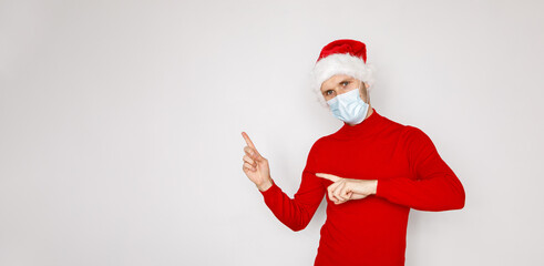 Fototapeta na wymiar Christmas during pandemic of COVID-19 Coronavirus. Man wearing surgical face mask and Santa hat. Male model wearing red sweater on white isolated background. Place for copy banner text seasonal Sales