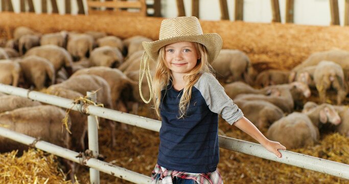 Portrait of Caucasian blonde pretty girl with fair hair standing at fence in stable and smiling to camera. Sheep flock on background. Cute little kid farmer in hat laughing in livestock barn.
