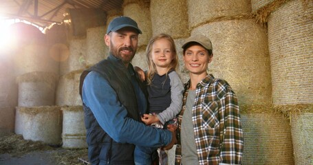 Portrait of happy Caucasian family of farmers with kid standing at barn with hay stocks and smiling...