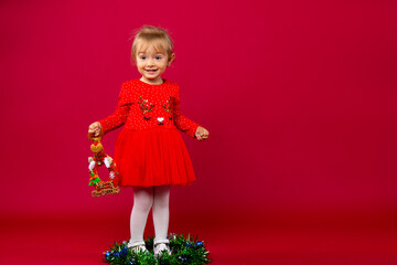 Blonde little girl in red Xmas dress with an reindeer decoration sitting in the middle of the christmas wreath against red background. Merry Christmas . Holiday concept