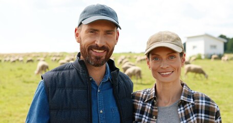 Portrait of happy young Caucasian couple of farmers in caps standing in hugs in green field and smiling to camera. Close up of wife and husband hugging at farm outdoor. Sheep flock on background.