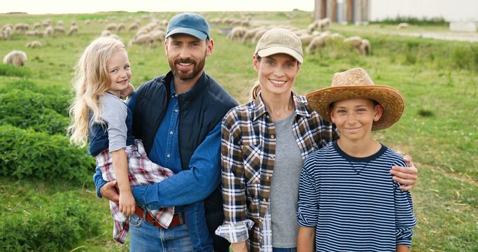 Portrait of Caucasian happy family with little kids standing at pasture with sheep flock on background and smiling to camera. Cheerful parents with small son and daughter in field at farm.