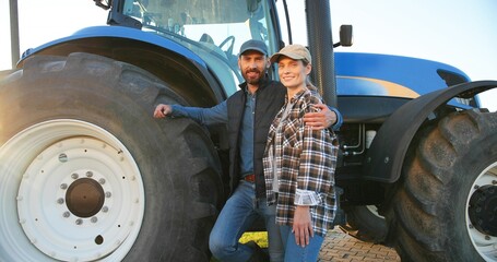 Portrait of Caucasian happy couple of married farmers standing in hugs at tractor, looking at each other and smiling to camera. Outdoor. Man hugging woman in field. Farming concept. Wife and husband. © VAKSMANV