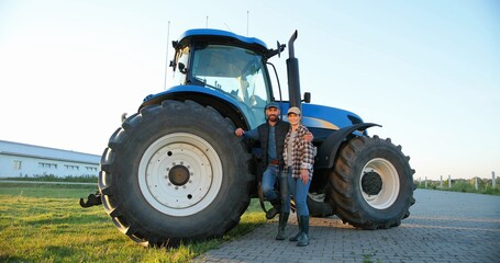 Portrait of Caucasian happy couple of married farmers standing in hugs at tractor, looking at each other and smiling to camera. Outdoor. Man hugging woman. Agricultural vehicle. Wife and husband.