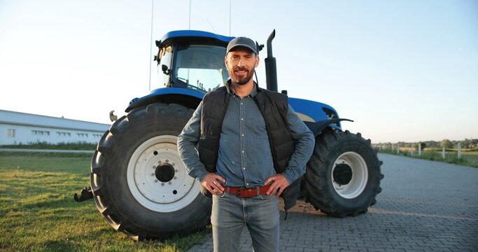 Portrait of young Caucasian handsome man farmer in cap standing on the road in field and smiling to camera. Big tractor on background. Male worker in agricultural farm. Agriculture farming.
