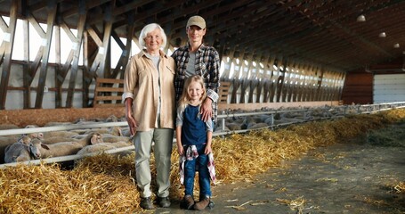 Portrait of Caucasian happy mother, old grandmother and cute small pretty girl in stable with sheep...
